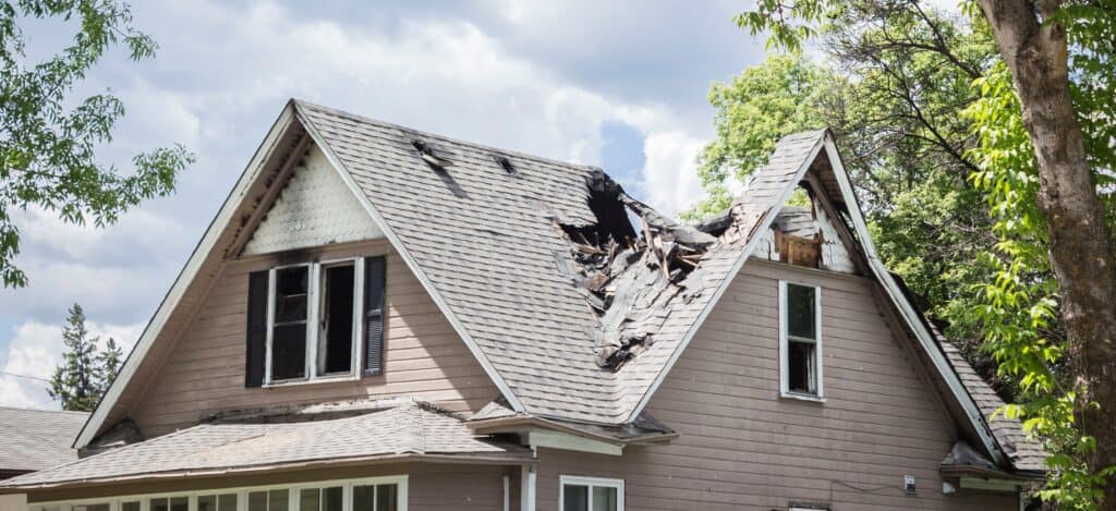 A large oak tree tossed by the winds of a summer storm falls onto and cuts through half of a house roof severely damaging it.